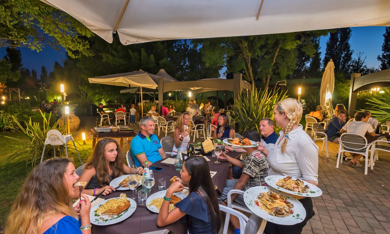 Diners eating meals served by a waitress under large umbrellas, surrounded by lush greenery and other dining tables at Isamar