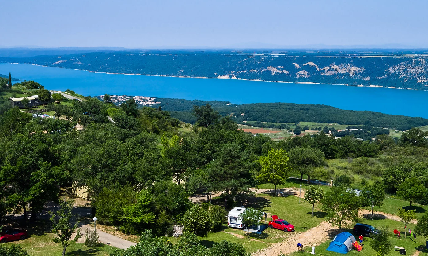 Campers setting up tents and vehicles in a lush, green park overlooking a blue lake with distant mountains at de l'Aigle