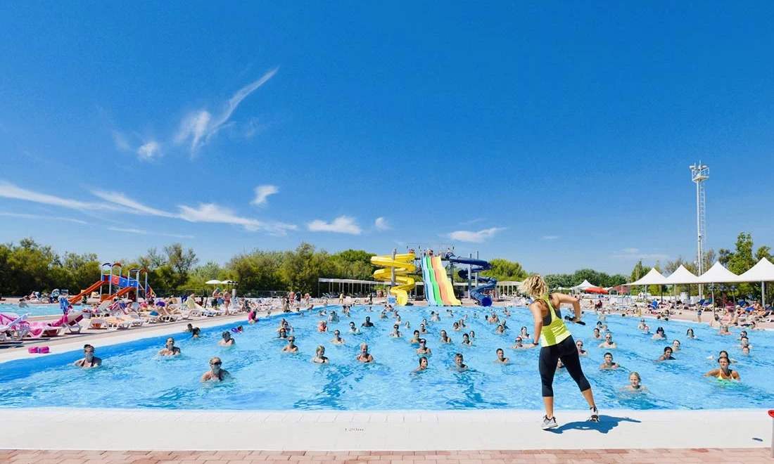 People swimming in a busy pool with slides while a woman instructs pool exercises, surrounded by trees and umbrellas at Isamar