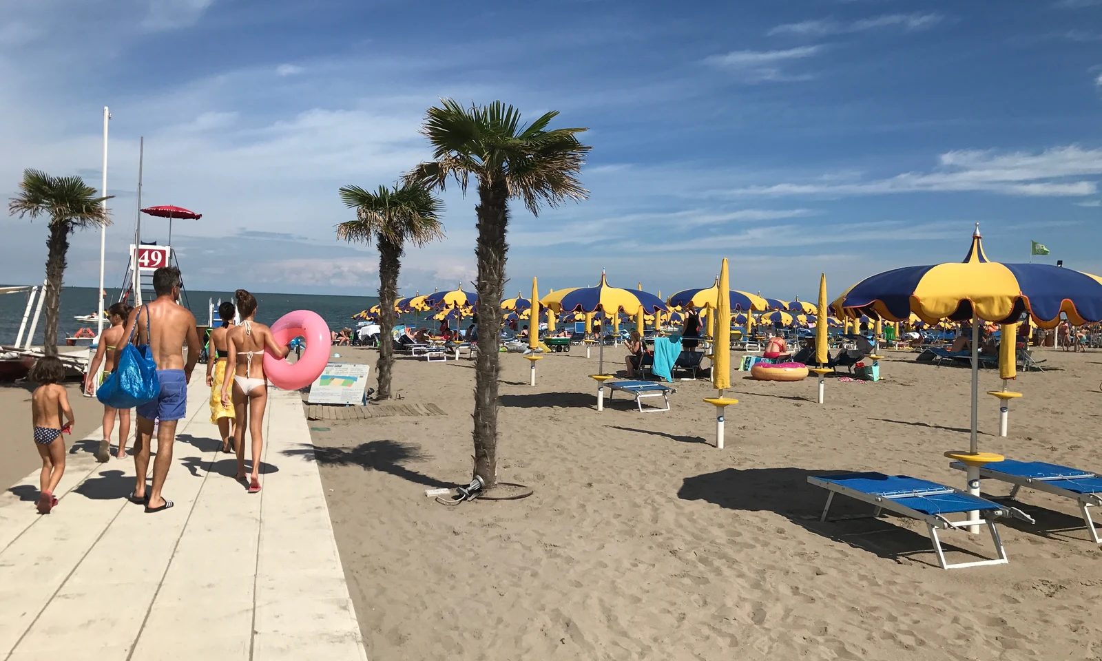 Family walking along a beach walkway, surrounded by palm trees, with yellow and blue umbrellas and sunbeds in the background at Isamar