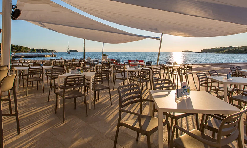 Empty outdoor dining tables and chairs overlook a calm waterfront with sailboats at sunset, shaded by large canopies at Orsera