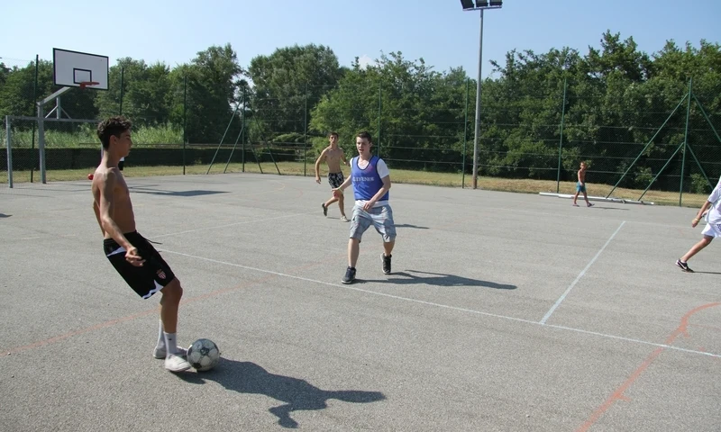 Young men playing soccer on an outdoor sports court surrounded by greenery and trees at Leï Suves