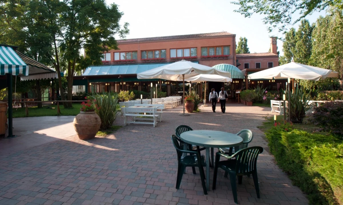 Table with four chairs on brick patio, surrounded by greenery; two people walking towards building with striped awnings at Isamar