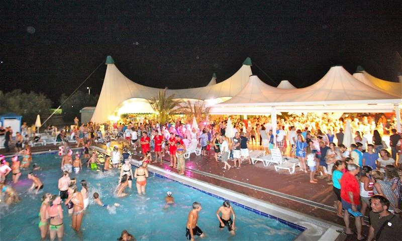 People swimming and socializing in a brightly lit pool area during a nighttime event, surrounded by tents and onlookers at Isamar