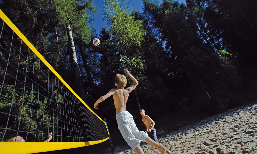 Man jumps to hit volleyball over net, surrounded by trees on sandy ground at Natterer See