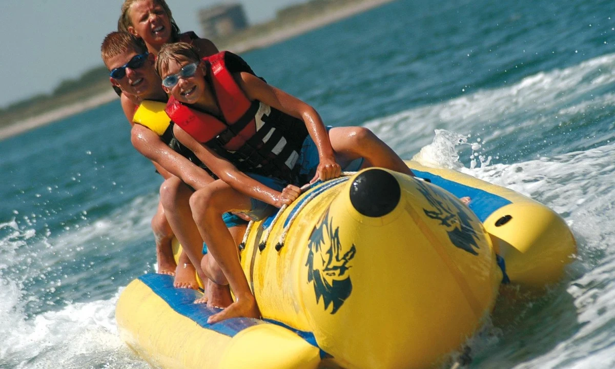 Three teenagers ride a yellow banana boat, skimming across the water near a sandy coastline at Isamar