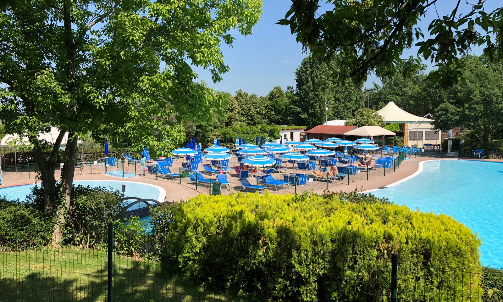 People lounging on sunbeds under blue umbrellas around a large swimming pool surrounded by greenery at Centro Vacanze San Marino