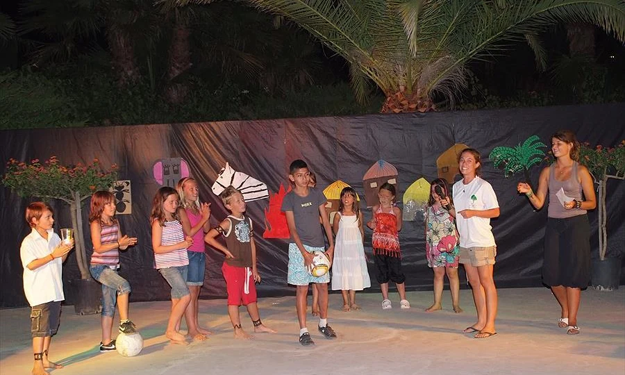 Children stand on stage holding props and performing a skit with painted backdrop and palm trees at Leï Suves