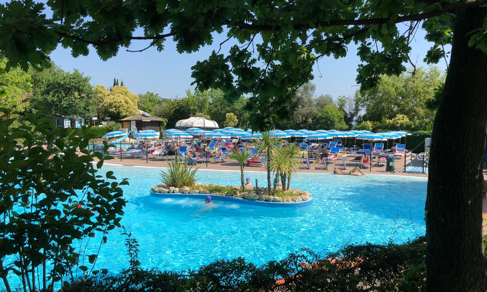 A pool with an island of plants is surrounded by people relaxing on sun loungers beneath umbrellas at Centro Vacanze San Marino