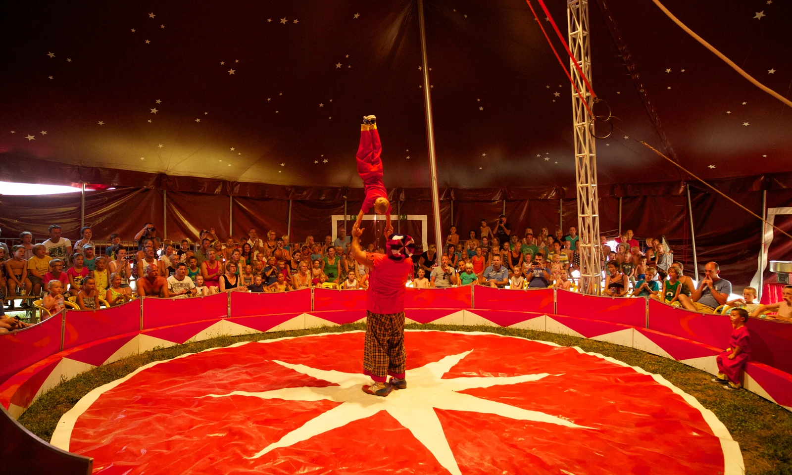 Circus performer balances a small child on one hand inside a tent, surrounded by an audience at Les Arches