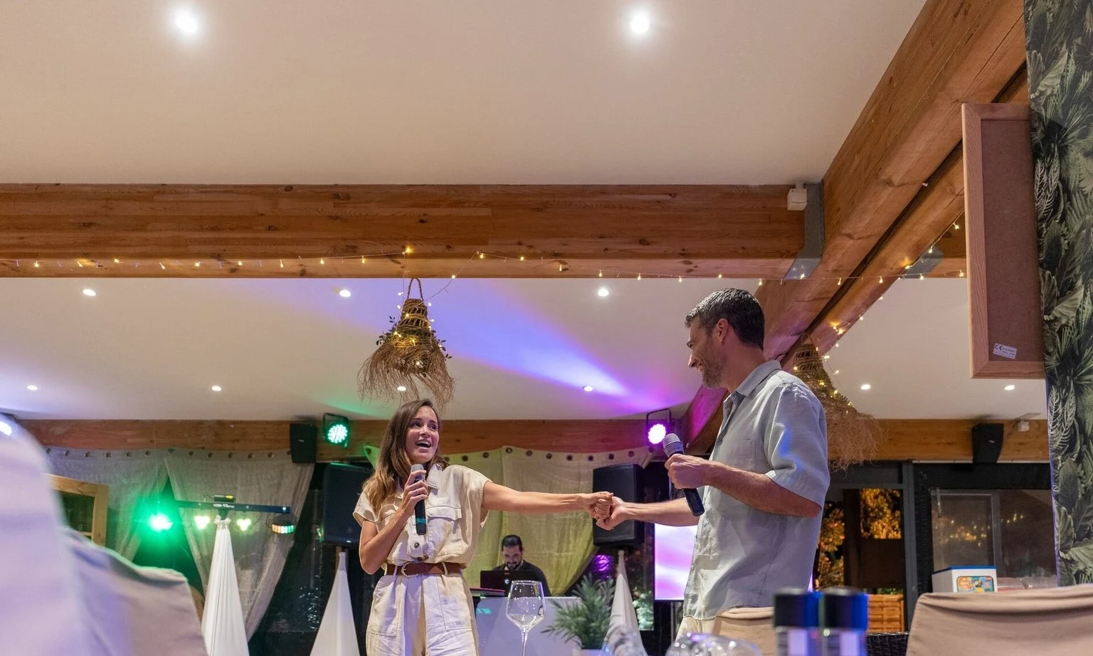 Two people smiling, holding microphones and hands, with festive lighting and decorations in an indoor setting at La Barque