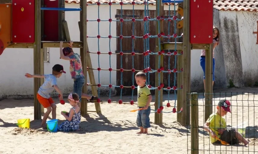 Children play on climbing structure and in sand; building with a tiled roof in the background at Les Amiaux