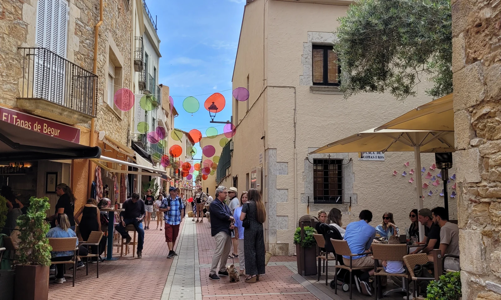 People dining outdoors and walking on a narrow, decorated street lined with stone buildings at Begur