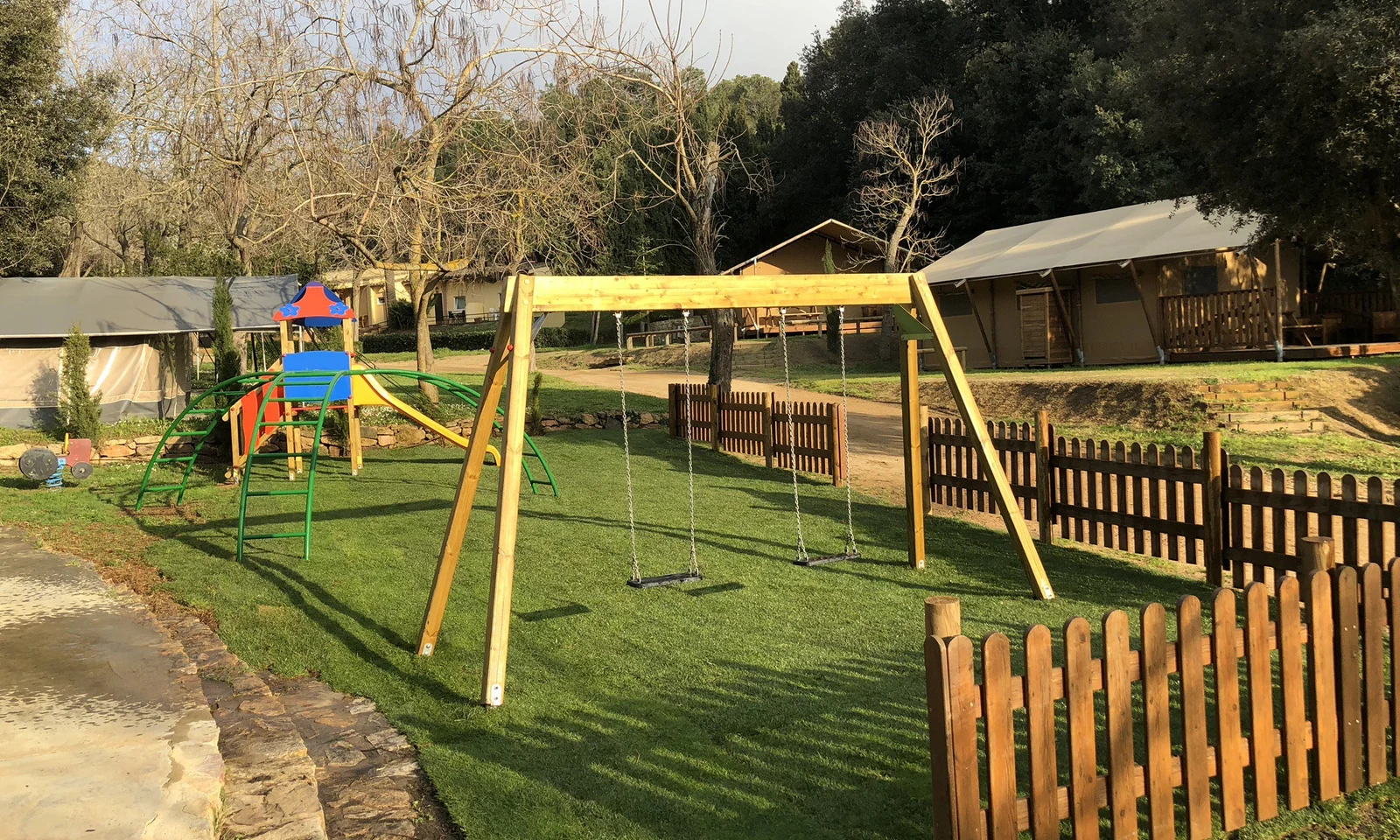 Children's playground with swings and climbing structure in a fenced grassy area, surrounded by tents and trees at Begur