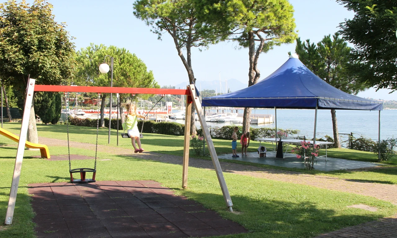 Child swinging in playground with trees, blue tent, and waterfront in the background at Front Lake Resort Le Corti del Lago