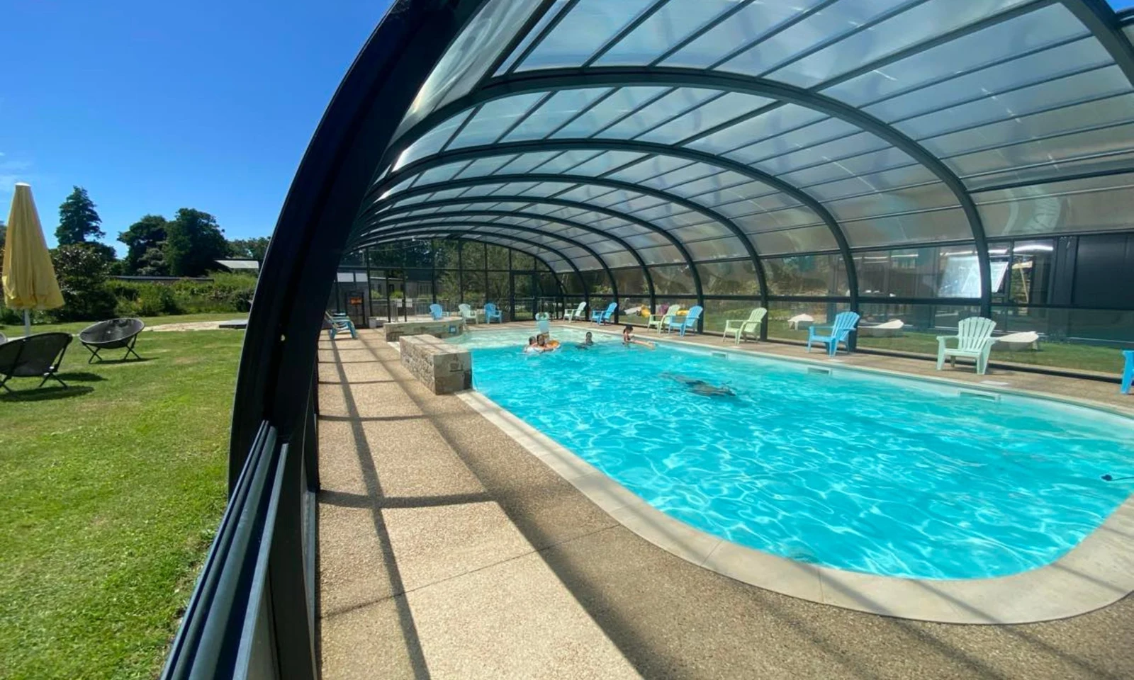 Enclosed swimming pool with people swimming, surrounded by lounge chairs and umbrellas, adjacent to a grassy area at Domaine de Mesqueau