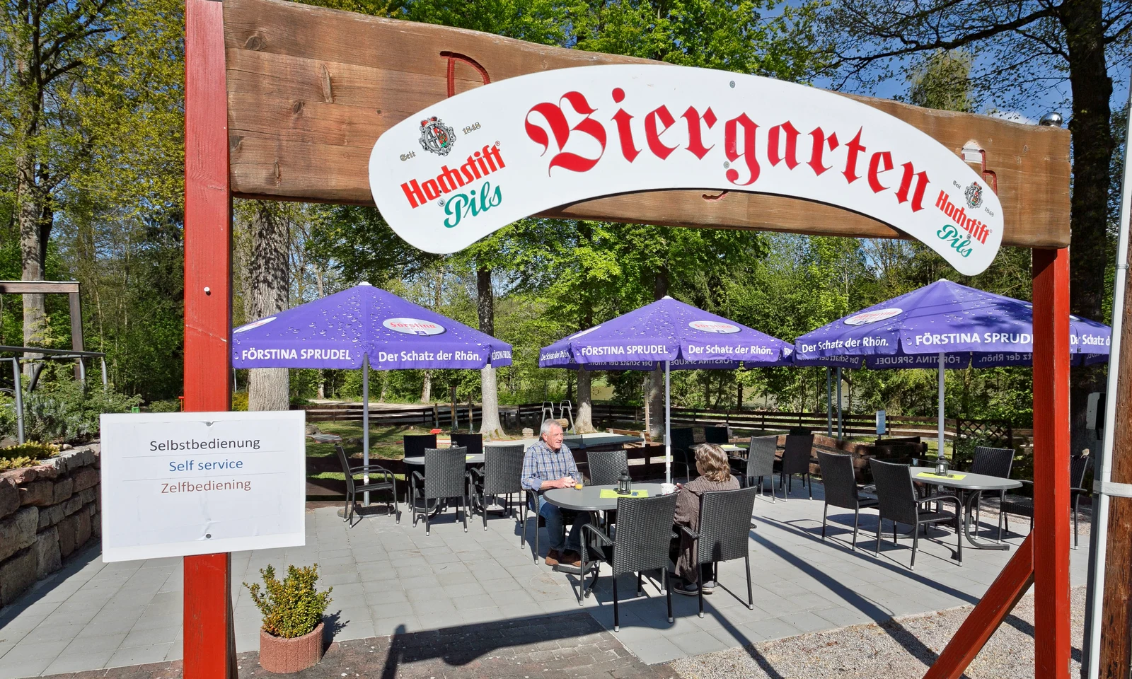 Outdoor seating area under purple umbrellas in beer garden, sunny day with trees in background at KNAUS Campingpark Hünfeld