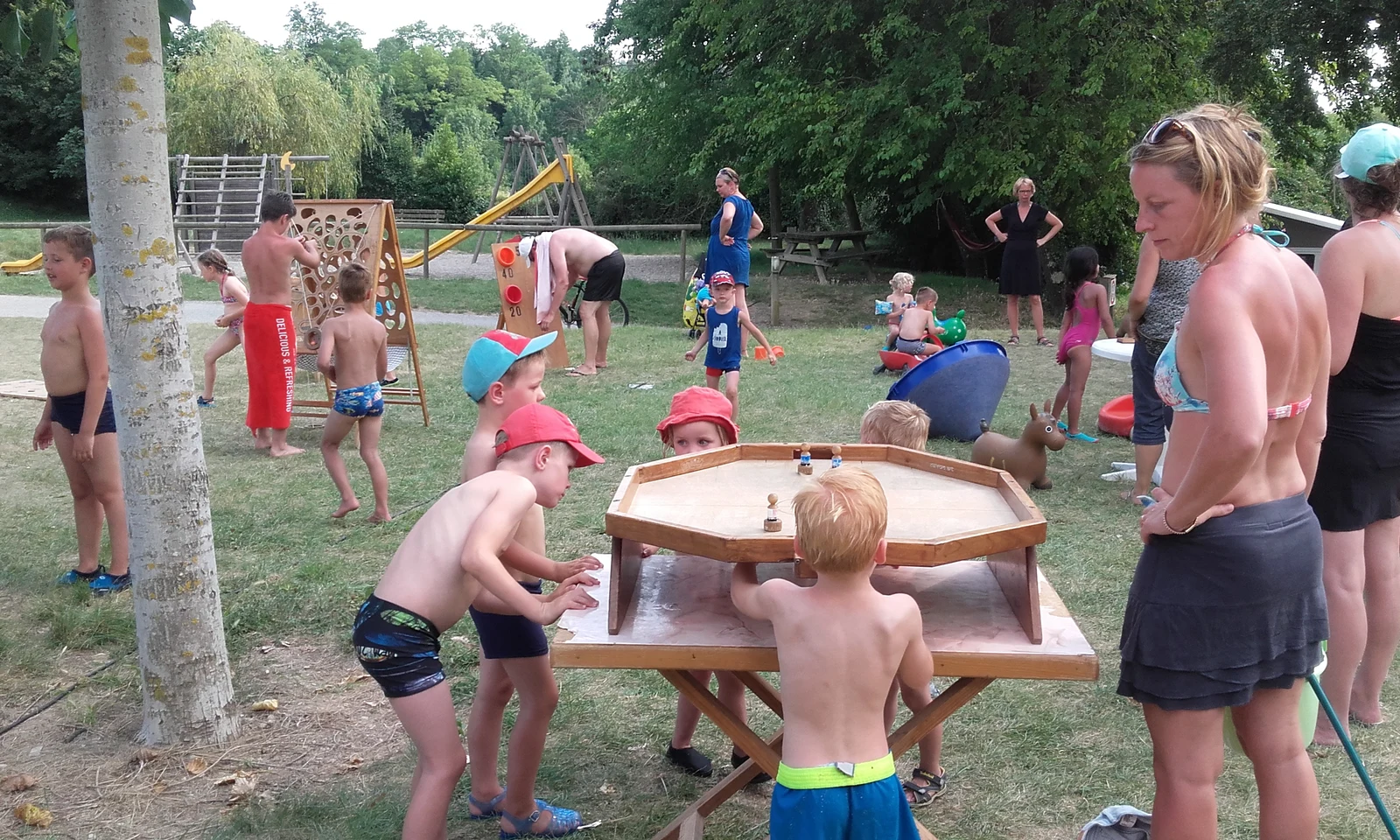 Children playing tabletop game with figurines outdoors, surrounded by other kids and adults engaged in various activities at Les Arches