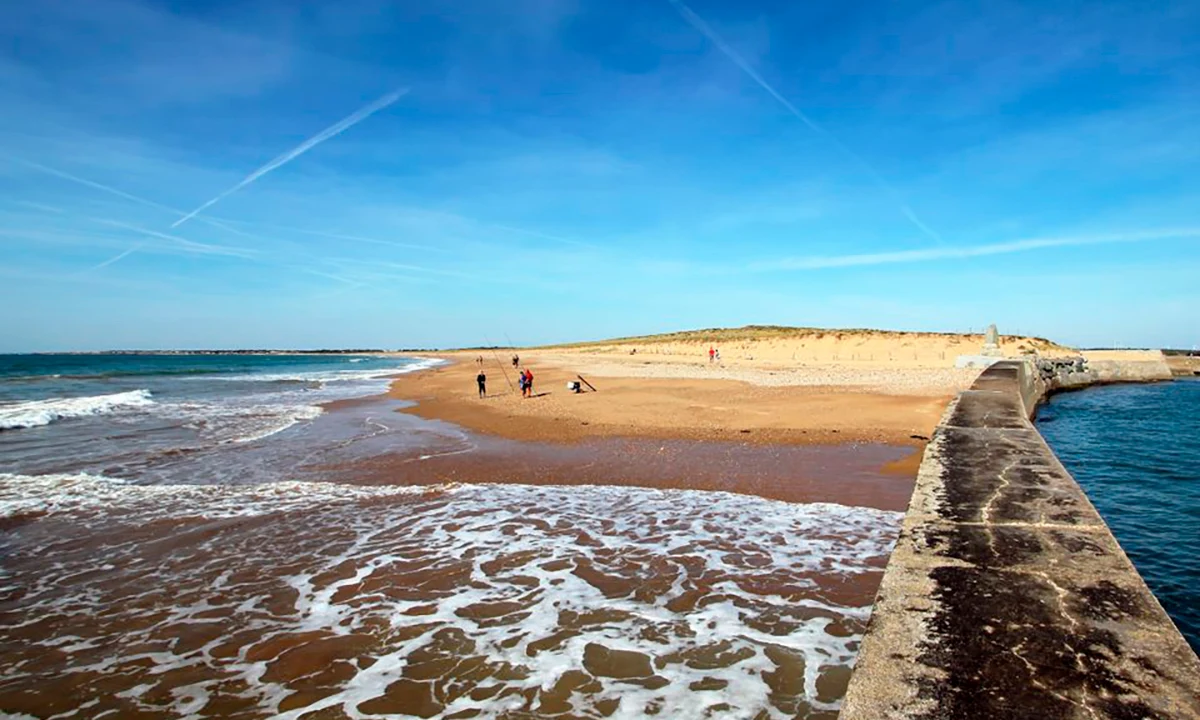 People fishing on a sandy beach beside a concrete pier, with gentle waves and a clear blue sky at Camping 2 Plages & Océan Village Corsaire