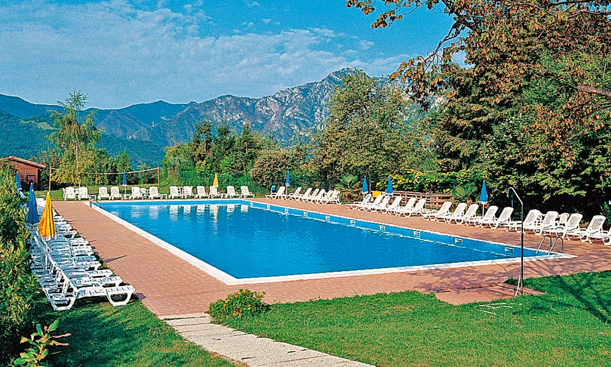 Pool surrounded by lounge chairs reflecting the mountain landscape with trees and blue sky at Residence Campi