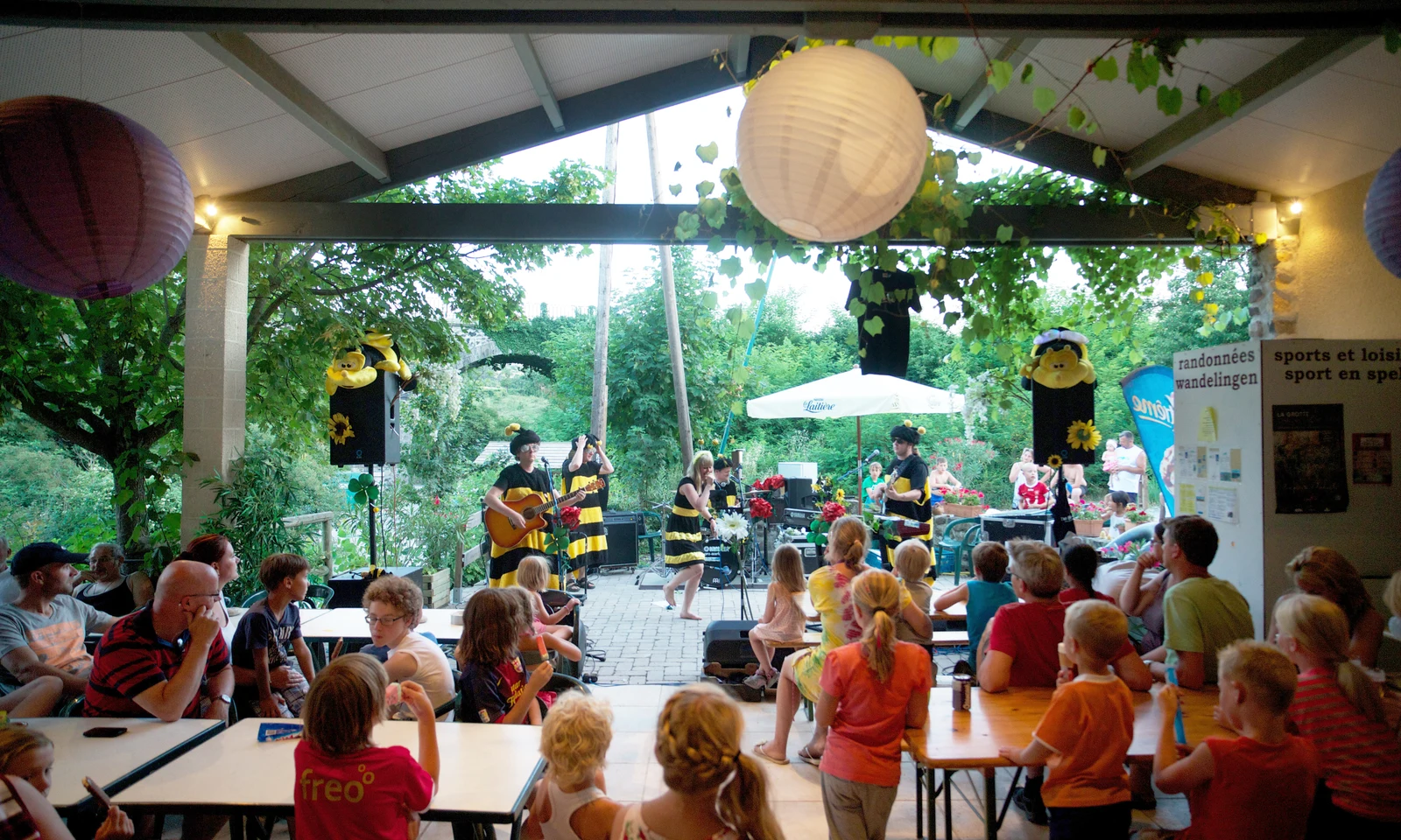 Performers in bee costumes singing, dancing on a stage outdoors, children and adults seated watching them at Les Arches