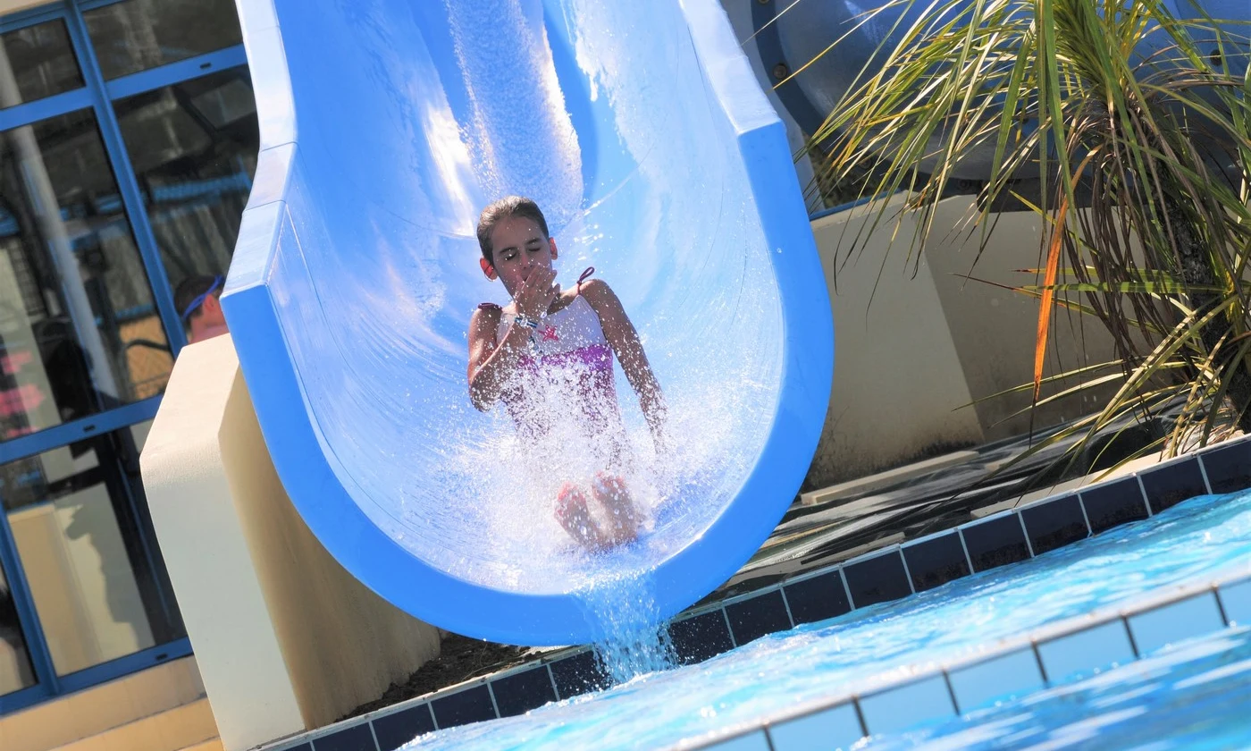 Girl in a pink swimsuit sliding down a blue waterslide into a pool, next to windows and tropical plants at Les Aventuriers de la Calypso