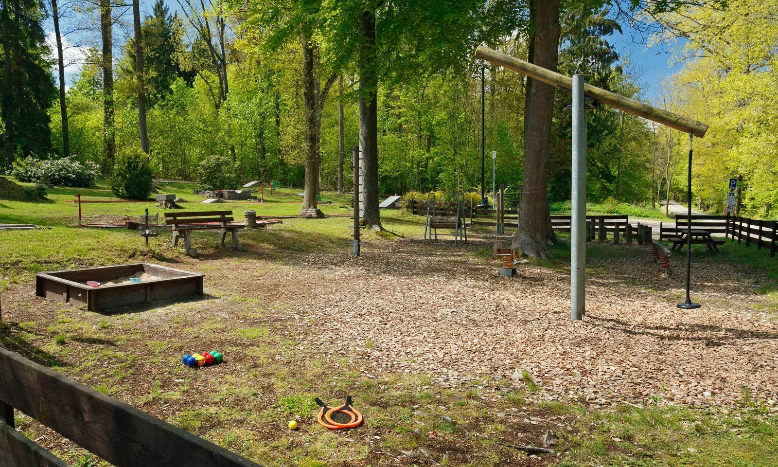 Playground equipment among trees and benches, surrounded by a grassy and wooded area at KNAUS Campingpark Hünfeld