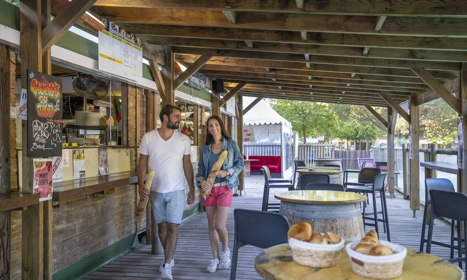 A man and woman hold baguettes while walking under a wooden awning beside a campsite café at Lac de Sanguinet