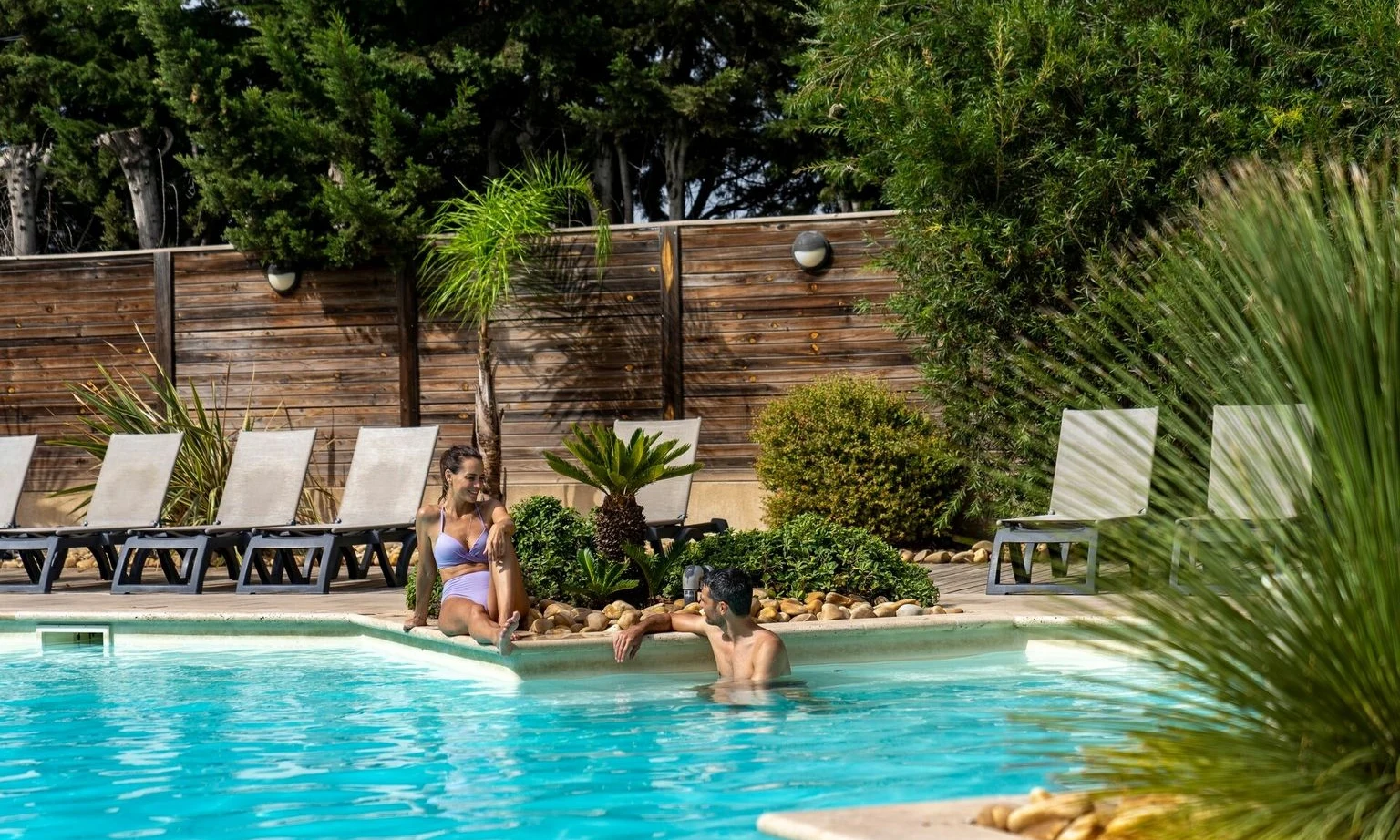 Two people relaxing by a swimming pool with lounge chairs, plants, and wooden fence in a sunny outdoor area at La Barque