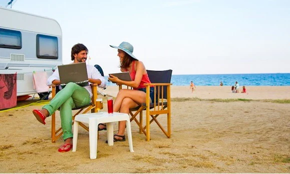 Two people sitting on chairs and using a laptop near a white camper, by the beach at Bon Repos