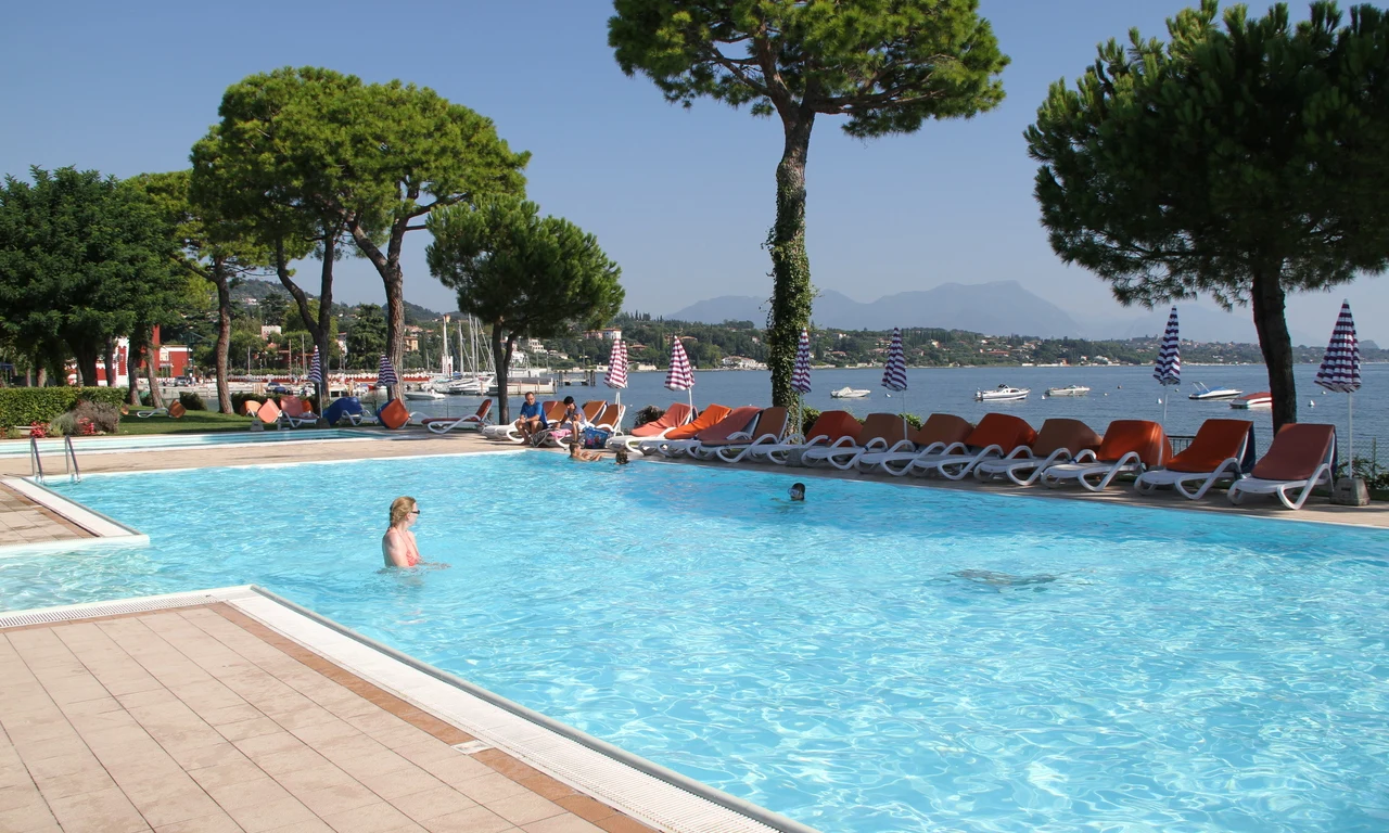 Swimming pool with loungers along the edge; people relax and swim; boats and trees in the background at Front Lake Resort Le Corti del Lago