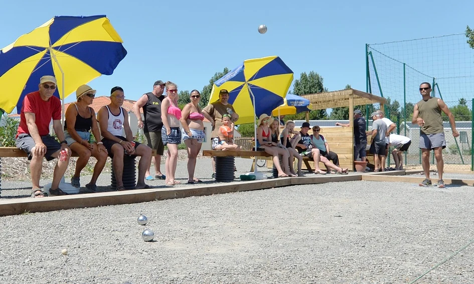 People watching a man play boules under large blue and yellow umbrellas at Les Aventuriers de la Calypso