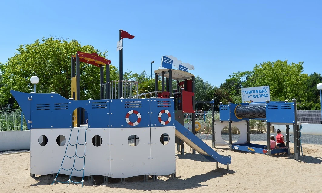 Blue and white ship-themed playground structure with slides and climbing nets in a sandy play area, surrounded by greenery at Les Aventuriers de la Calypso