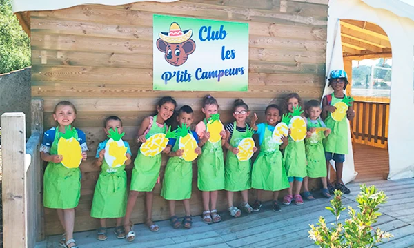 Children wearing green aprons hold pineapple cutouts, standing in front of a wooden structure at Les Aventuriers de la Calypso