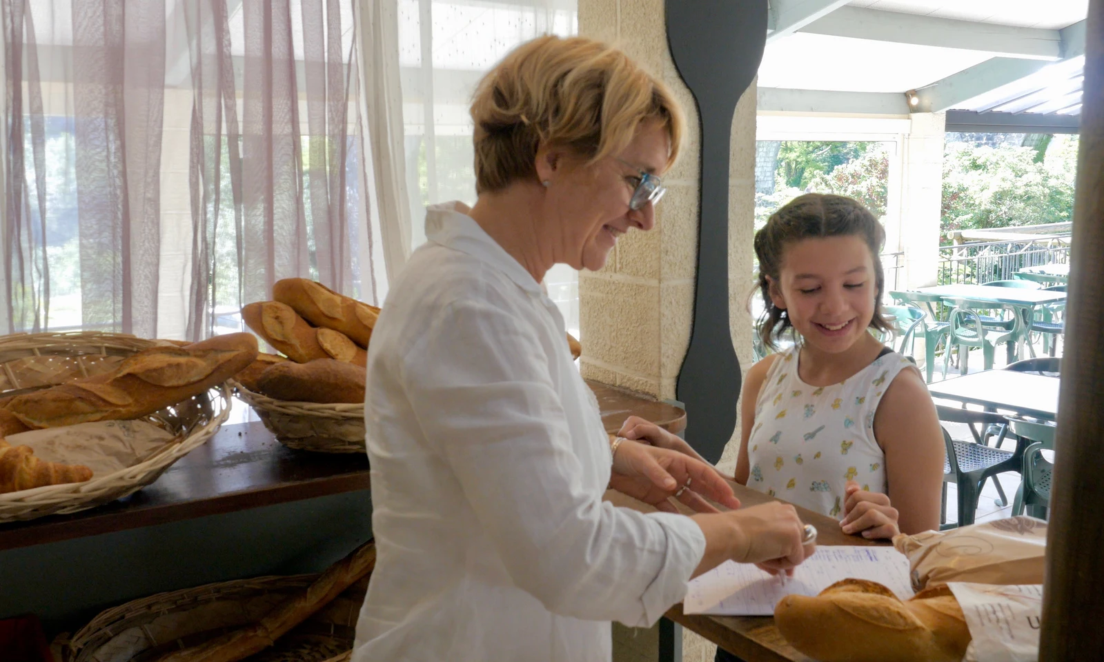 Person smiling, handing bread to child inside a well-lit café with bread baskets on the counter at Les Arches