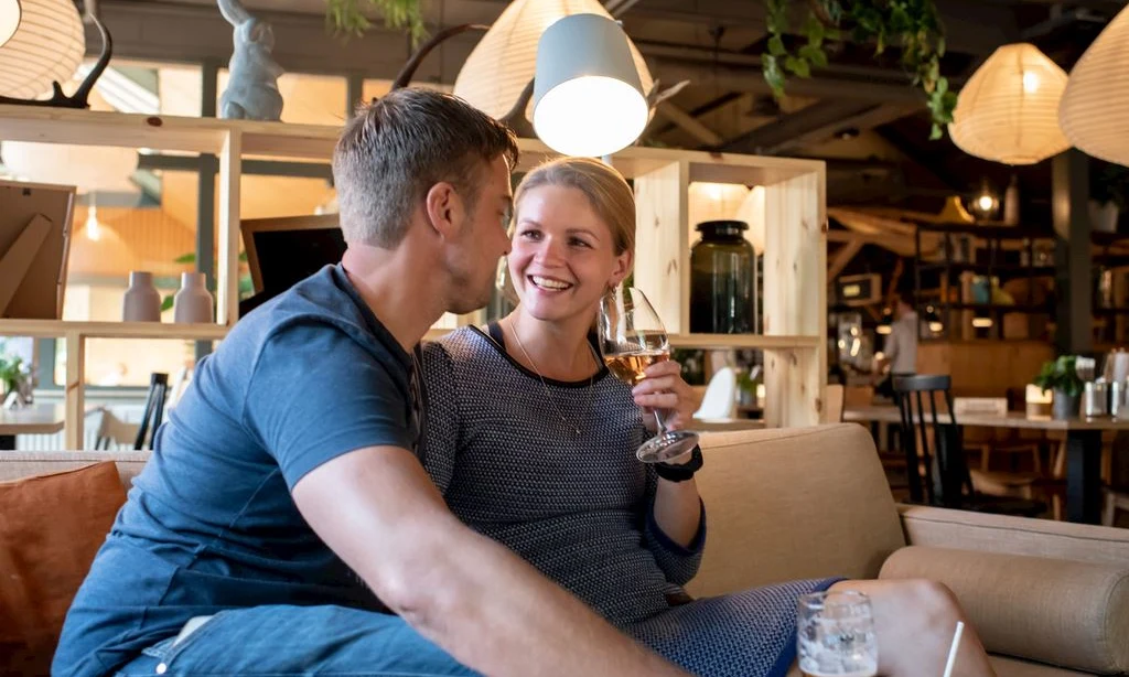 Couple sitting on a sofa, smiling and holding drinks, in a cozy, warmly-lit indoor setting at Landal Rabbit Hill