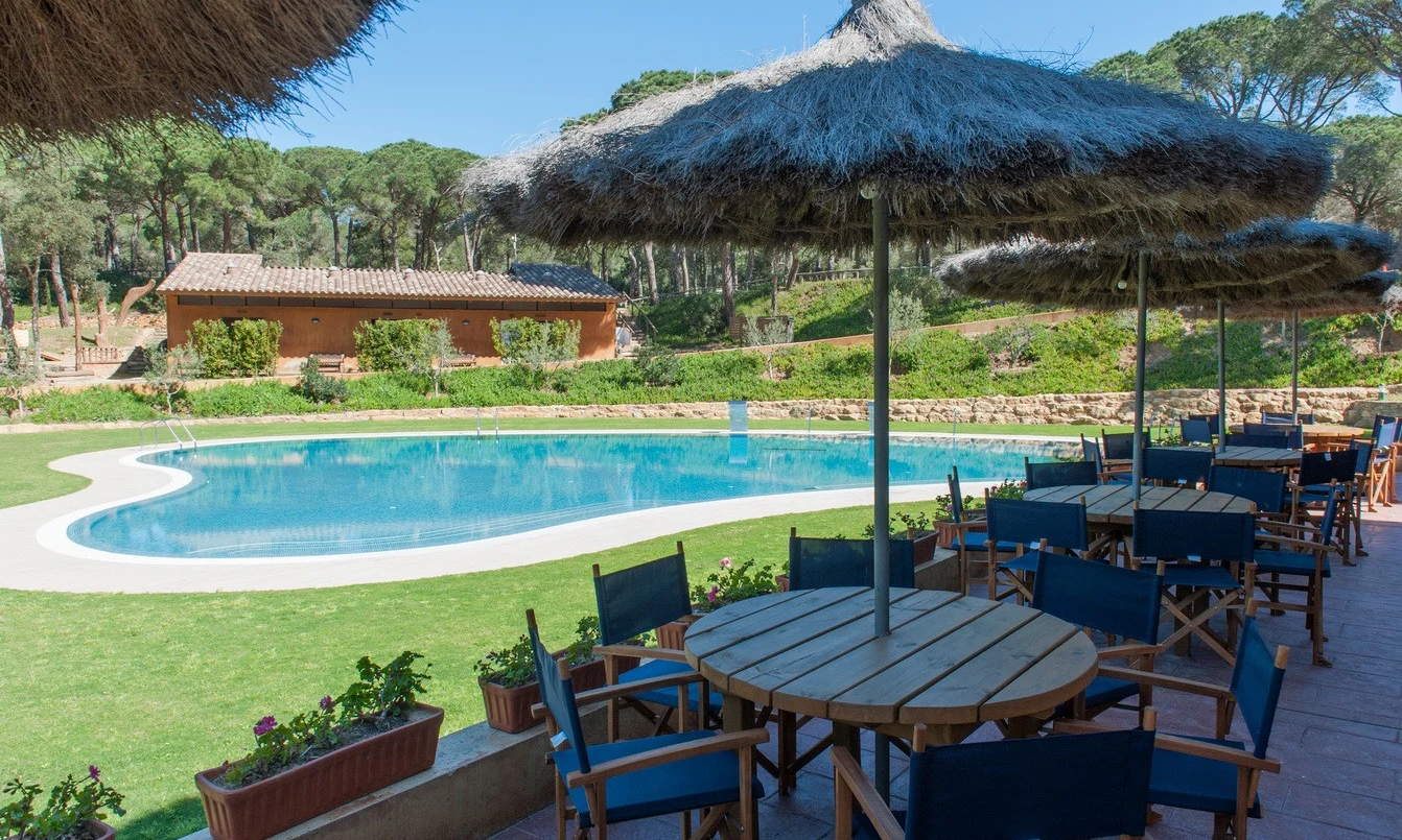 Wooden tables with blue chairs sit under thatched umbrellas near a pool, surrounded by greenery at Begur