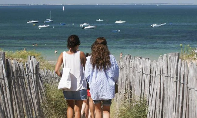 People walking down a sandy path between wooden fences towards a beach with boats on water at Seasonova Ile de Ré