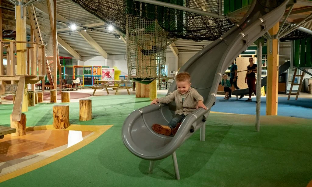 Child sliding down a grey slide, surrounded by indoor play area with wooden structures and nets at Landal Rabbit Hill