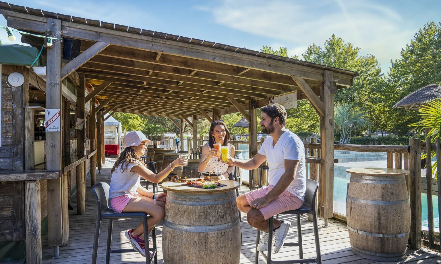 Three people enjoying drinks together outdoors at a rustic bar with a wooden roof near a pool at Lac de Sanguinet