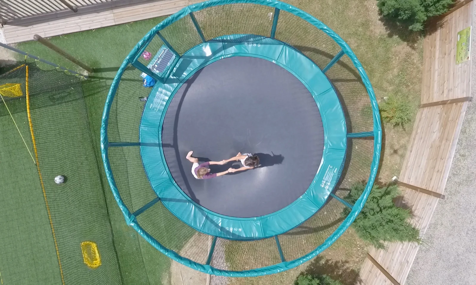 Trampoline in use by two children holding hands, situated in a yard surrounded by netting and wooden fencing at Les Arches