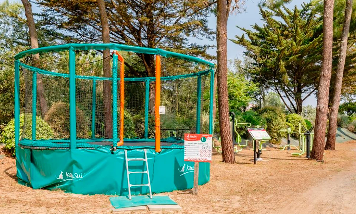 Trampoline with safety nets stands in a wooded camping area, next to outdoor gym equipment at Camping 2 Plages & Océan Village Corsaire