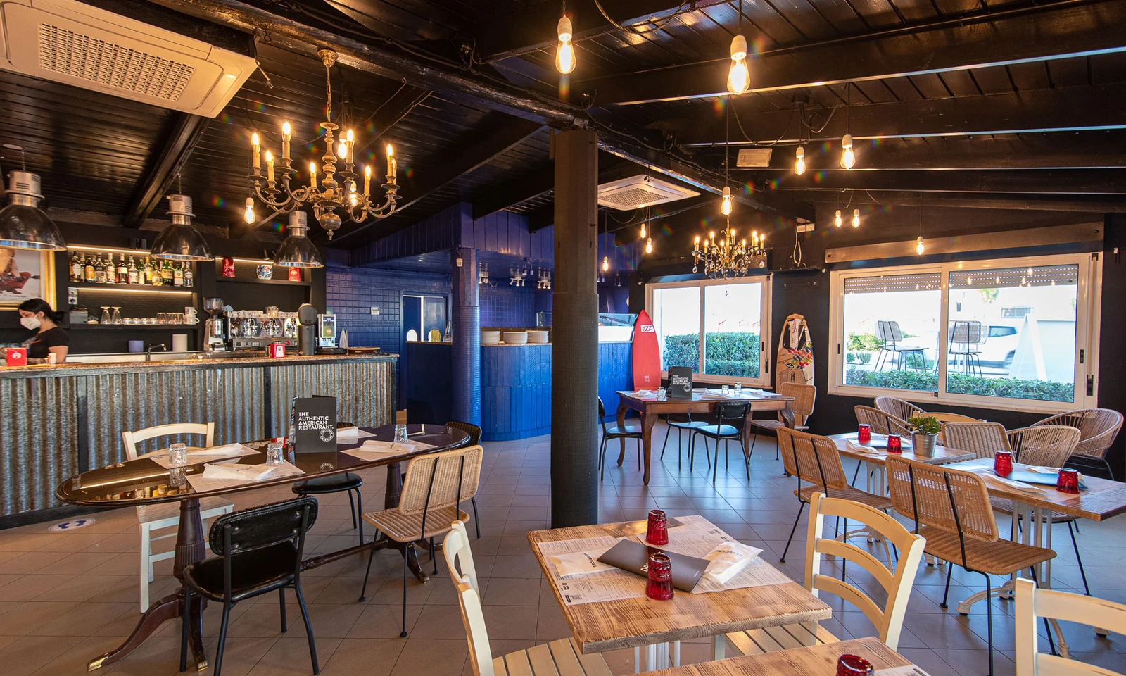 Restaurant interior features tables and chairs arranged under chandeliers, with a bar staffed by a masked employee at International Riccione Family Camping Village