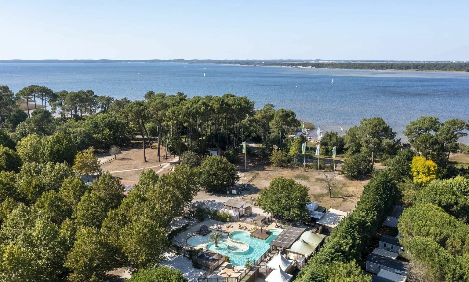 Swimming pool with surrounding cabins and greenery, set beside a lake with trees and sailboats in the background at Lac de Sanguinet