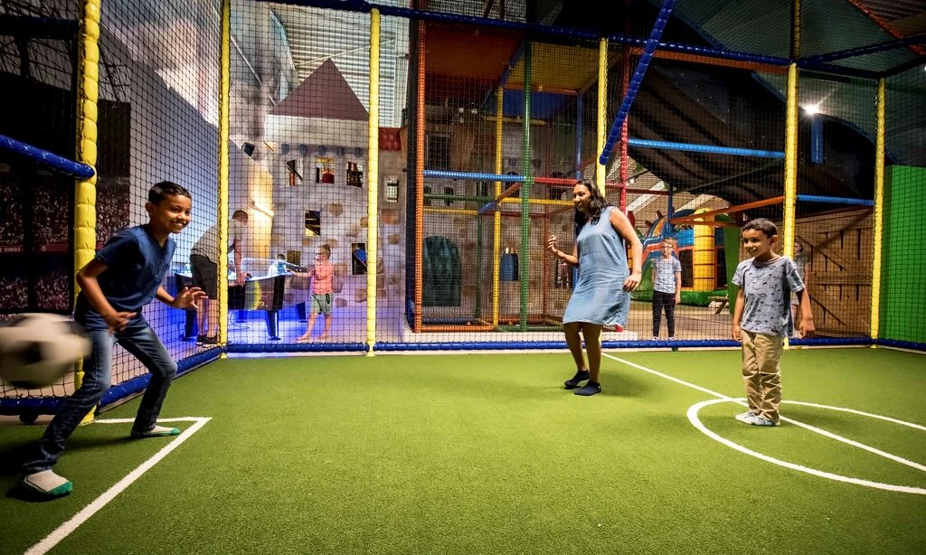 Children and adults playing soccer indoors on artificial turf, enclosed by colorful safety nets at Landal Rabbit Hill