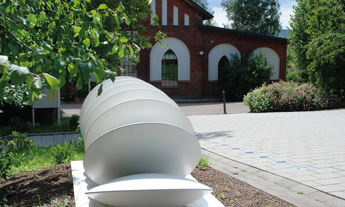 White cylindrical sculpture resting on a paved area, surrounded by green shrubs and trees with a brick building in the background at KNAUS Campingpark Hünfeld