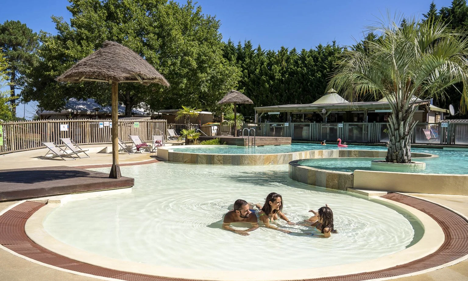 Family playing in a shallow pool surrounded by umbrellas and deck chairs, near lush trees at Lac de Sanguinet