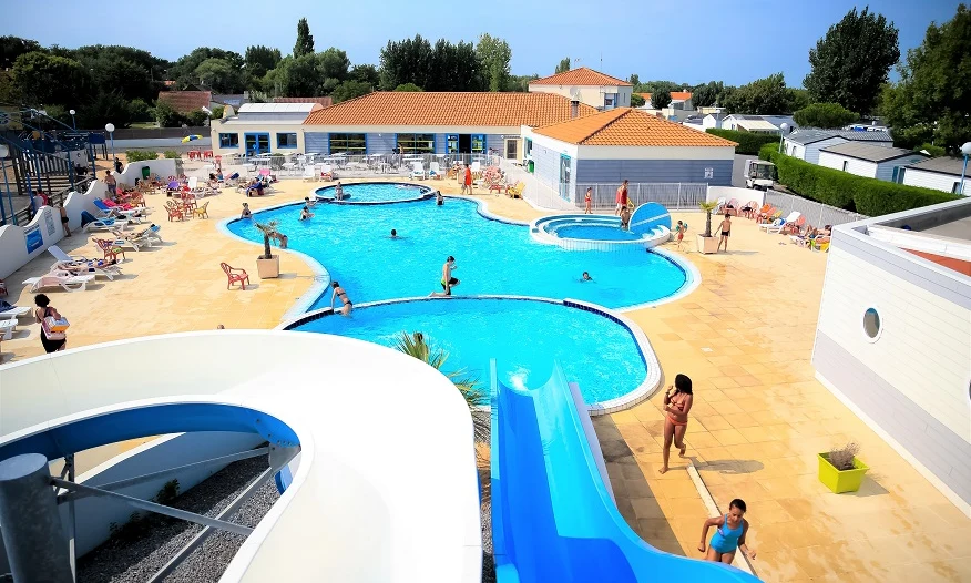 Outdoor swimming pool with water slides and people swimming and sunbathing, surrounded by buildings and greenery at Les Aventuriers de la Calypso