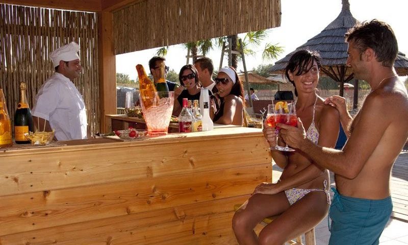 People are socializing and enjoying drinks at a wooden outdoor bar with a bartender under a hut near the beach at Oasi