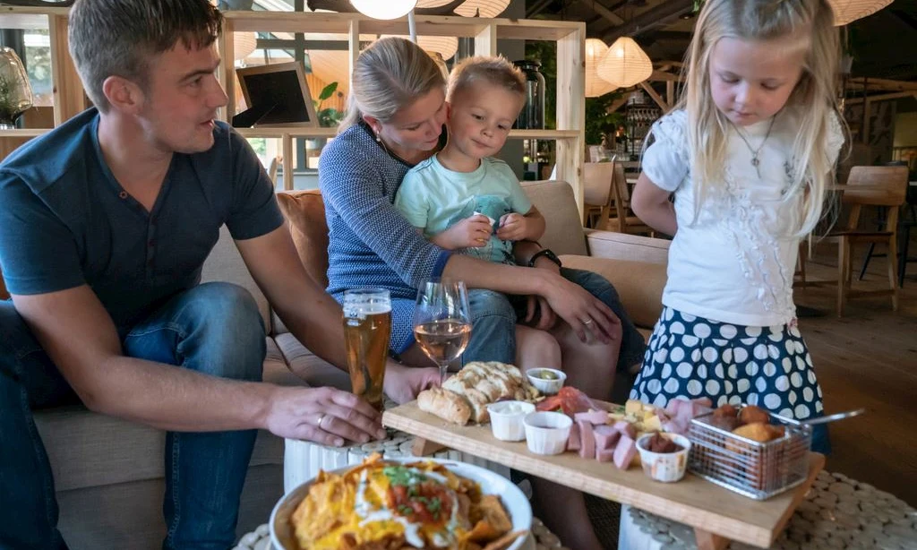 Family seated on a couch enjoying appetizers together in a cozy, warmly lit dining area with wooden accents at Landal Rabbit Hill